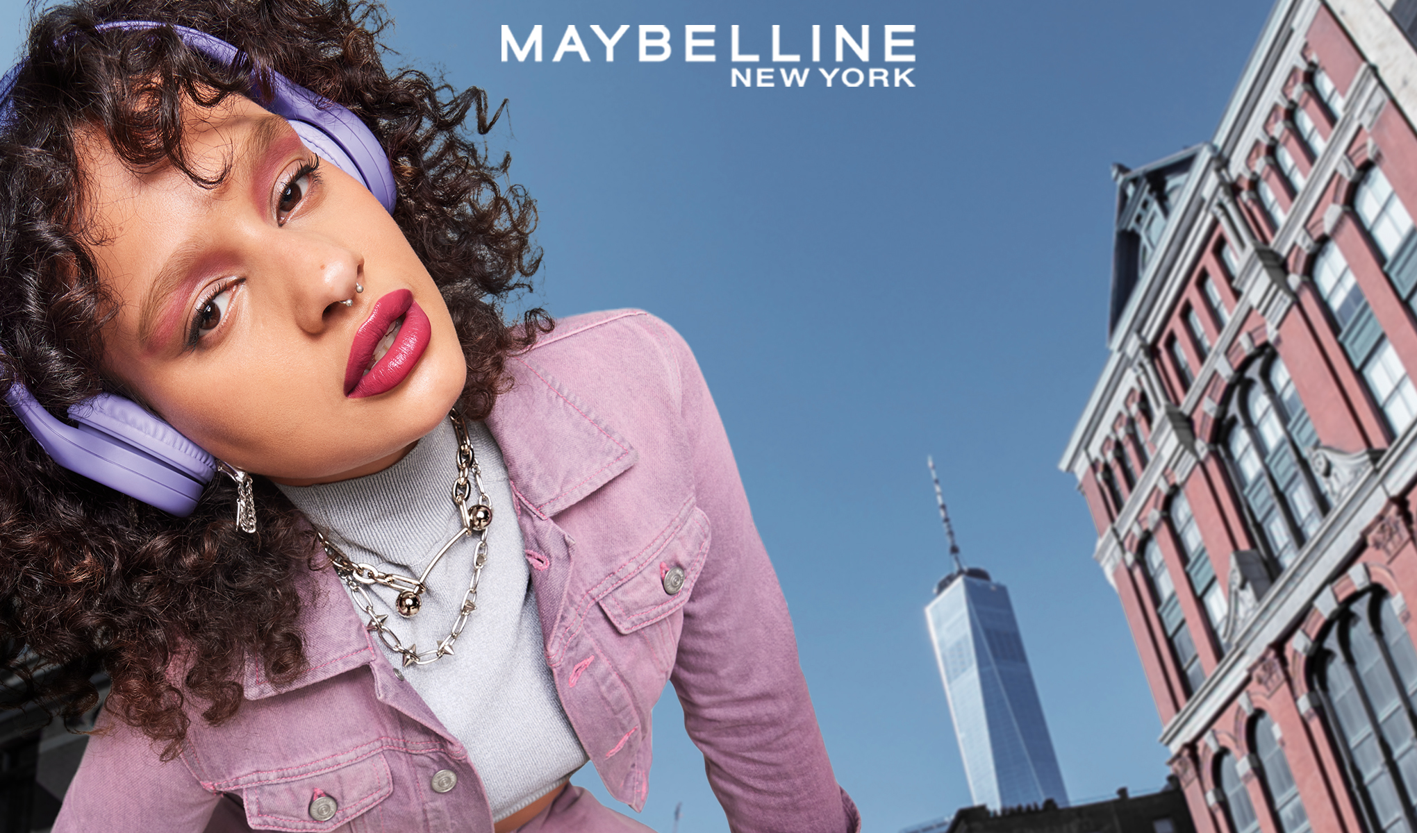 Kiharatukkainen nainen, jolla on violetit kuulokkeet ja vaaleanpunainen takki. Hänen yläpuolellaan on Maybelline New York valkoisella. Taustalla on sininen taivas ja rakennus.